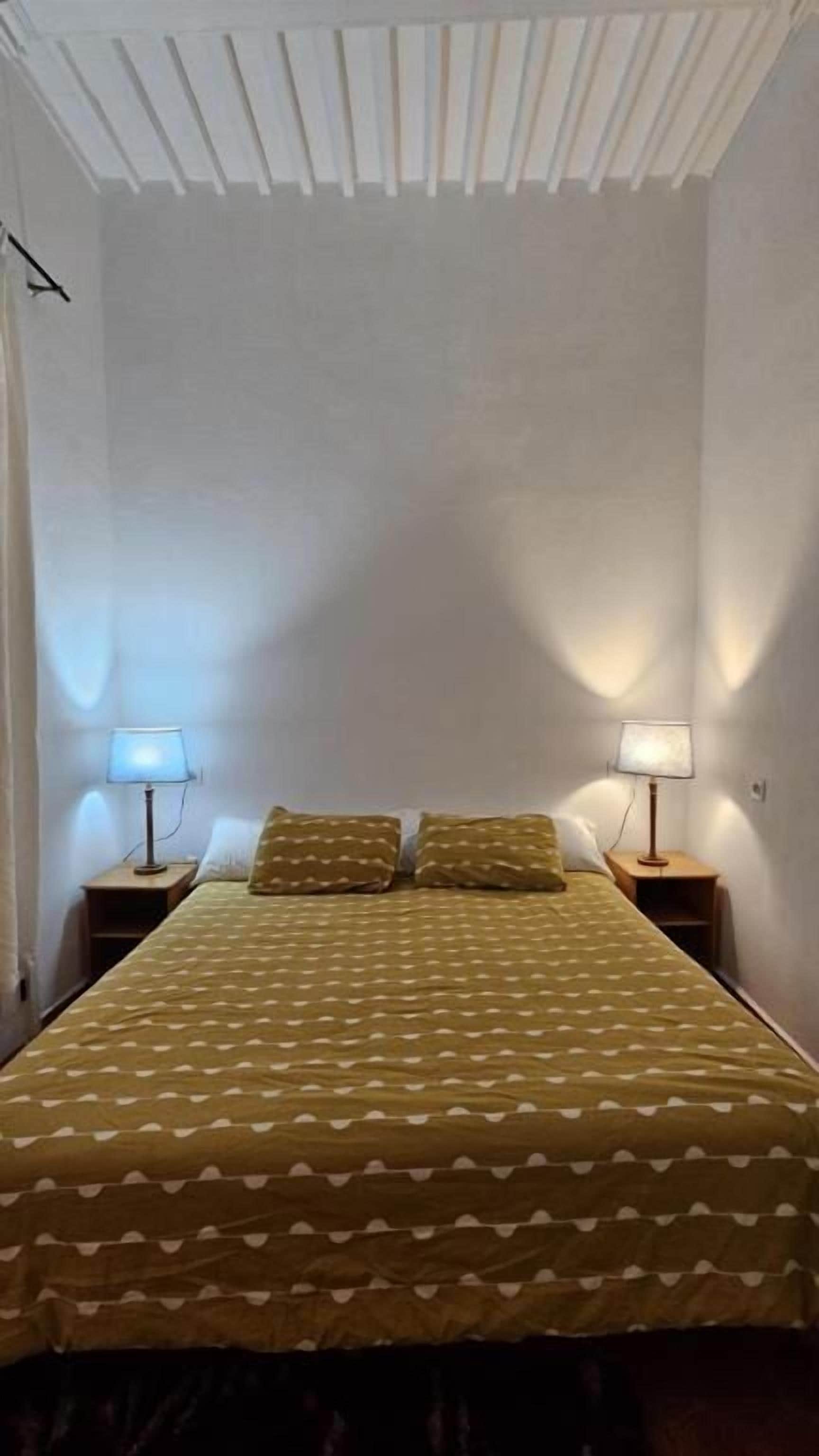 A small, single-bed room with a patterned bedspread, two bedside lamps, and a sloped ceiling.