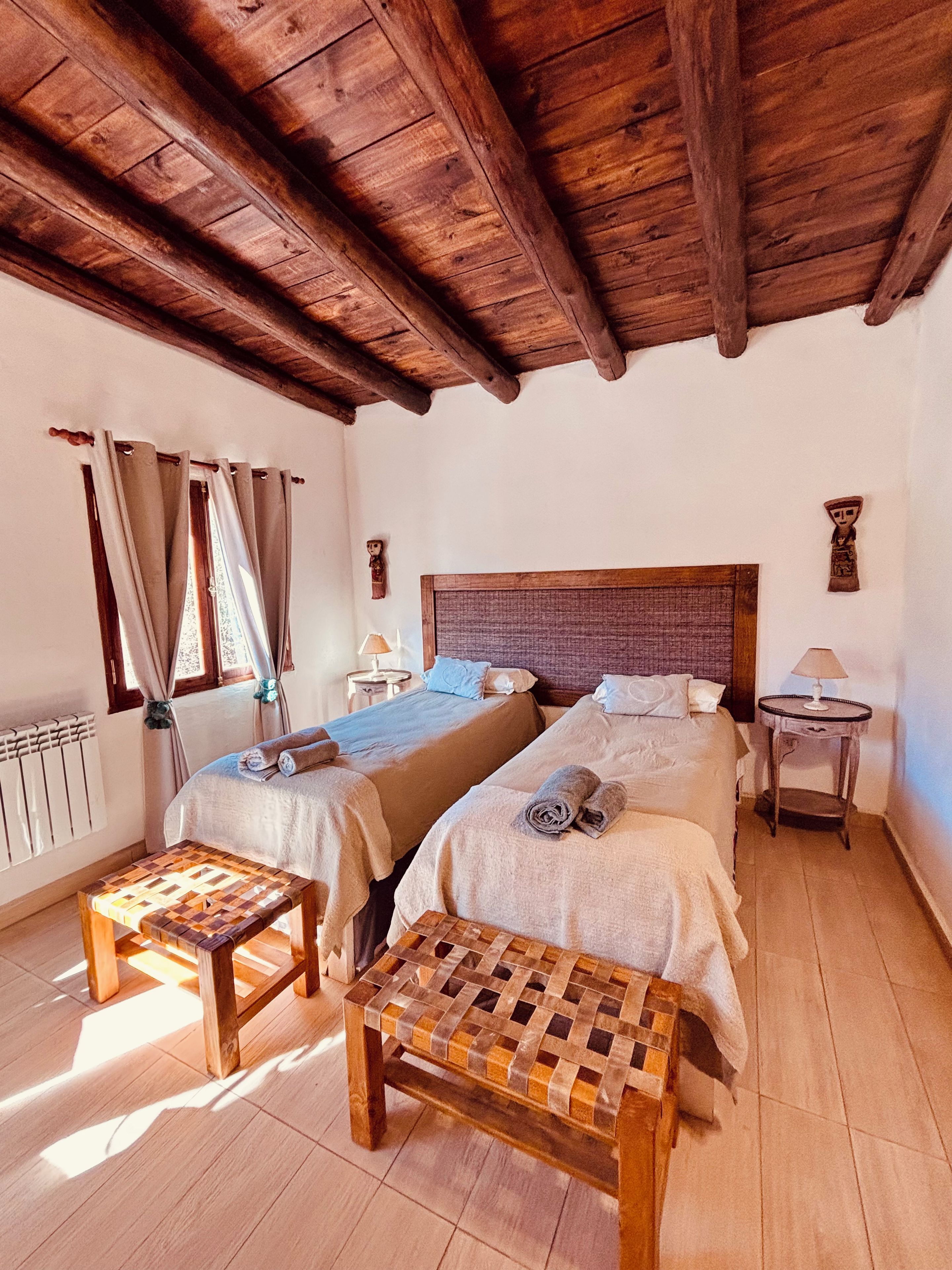 Una habitación con dos camas, un banco de madera, una mesa y un ventanal con cortinas.