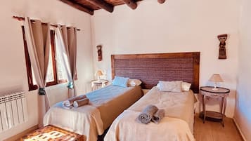 Una habitación con dos camas, un banco de madera, una mesa y un ventanal con cortinas.