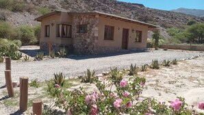 Una casa de piedra con camino de grava, rodeada de vegetación desértica y montañas.