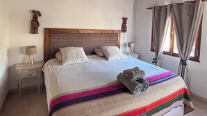 Un dormitorio con techo de madera, cama con ropa blanca, manta a rayas de colores y ventana con cortinas.