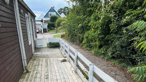Une terrasse en bois avec une rambarde blanche, entourée de verdure et bordée d’un bâtiment.