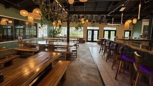 L’intérieur d’un restaurant est aménagé avec des tables et des bancs en bois, des suspensions lumineuses et de grandes fenêtres qui laissent entrer la lumière naturelle.