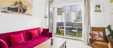 Ein Wohnzimmer mit einem leuchtend roten Sofa, einem Holztisch und einem braunen Sessel mit einem blumigen Kissen. An der Wand hängt ein Gemälde von einem Schiff und einem Seemann, und durch das Fenster ist ein Garten und ein Gebäude im Freien zu sehen.