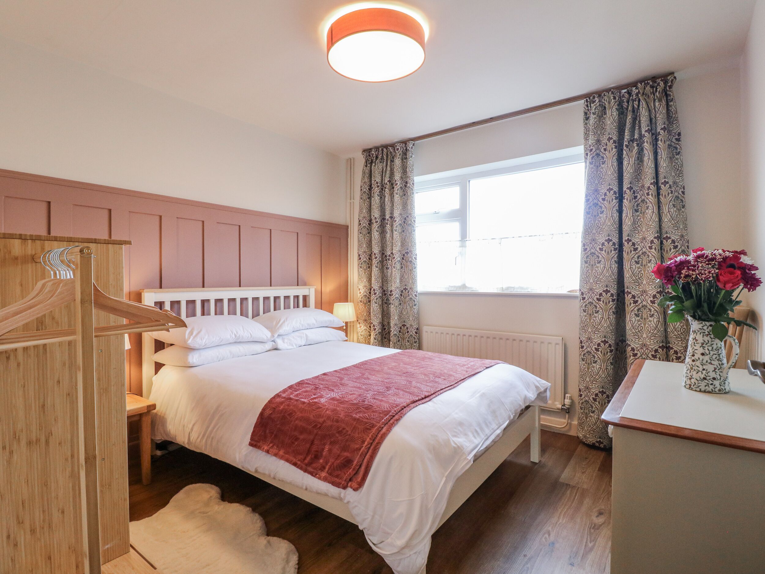 A bedroom with a bed, a wooden wardrobe, a radiator, a window with curtains, and a vase with flowers on a bedside table.