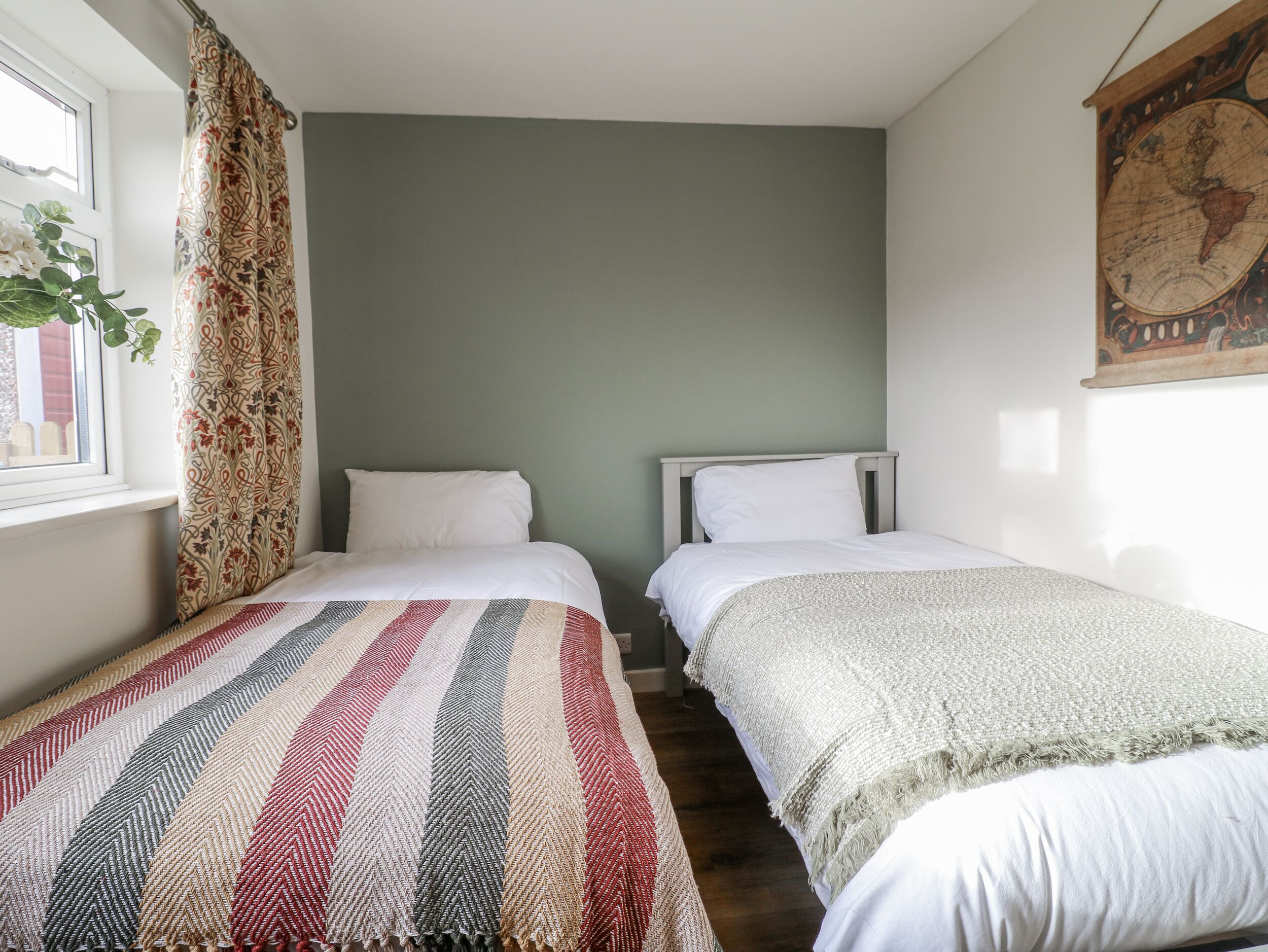 A room with two beds, a window with curtains, a map on the wall, and a plant by the window.