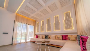 A modern living room with a sofa, a round coffee table, and a large window with decorative lighting. - Villa Lumeria (Marrakech)