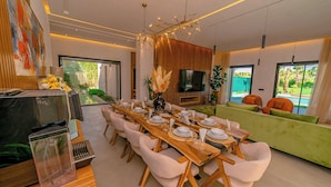 A modern dining area with a long wooden table set for six, surrounded by plush chairs. A television is mounted on the wall, and there is a green sofa with orange cushions in the background. - Villa Lumeria (Marrakech)