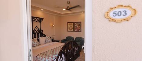 A bedroom with a bed, a ceiling fan, a wall-mounted number plate, and two chairs.