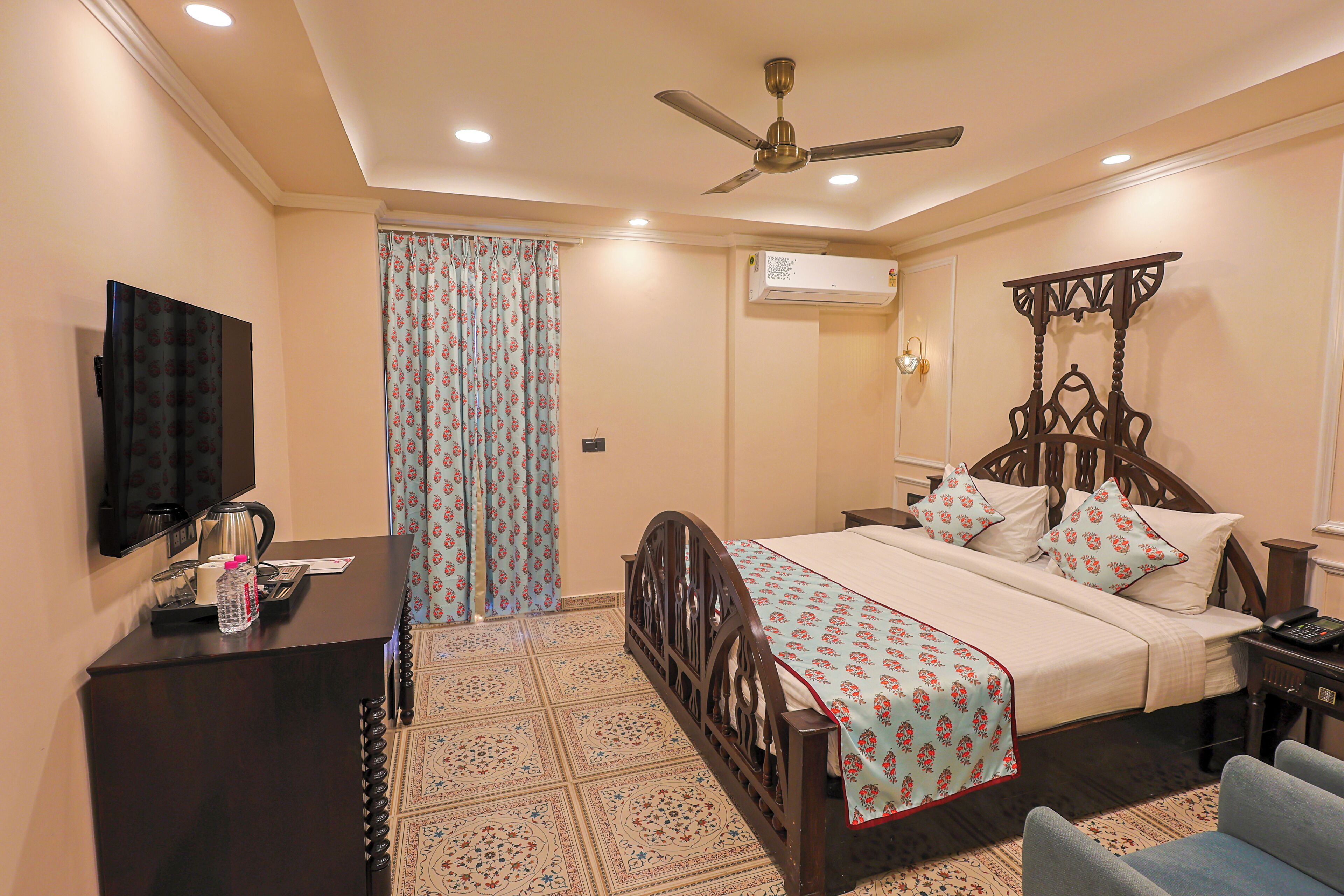 A hotel room with a bed, a desk with a television, a ceiling fan, an air conditioning unit, and a patterned floor.