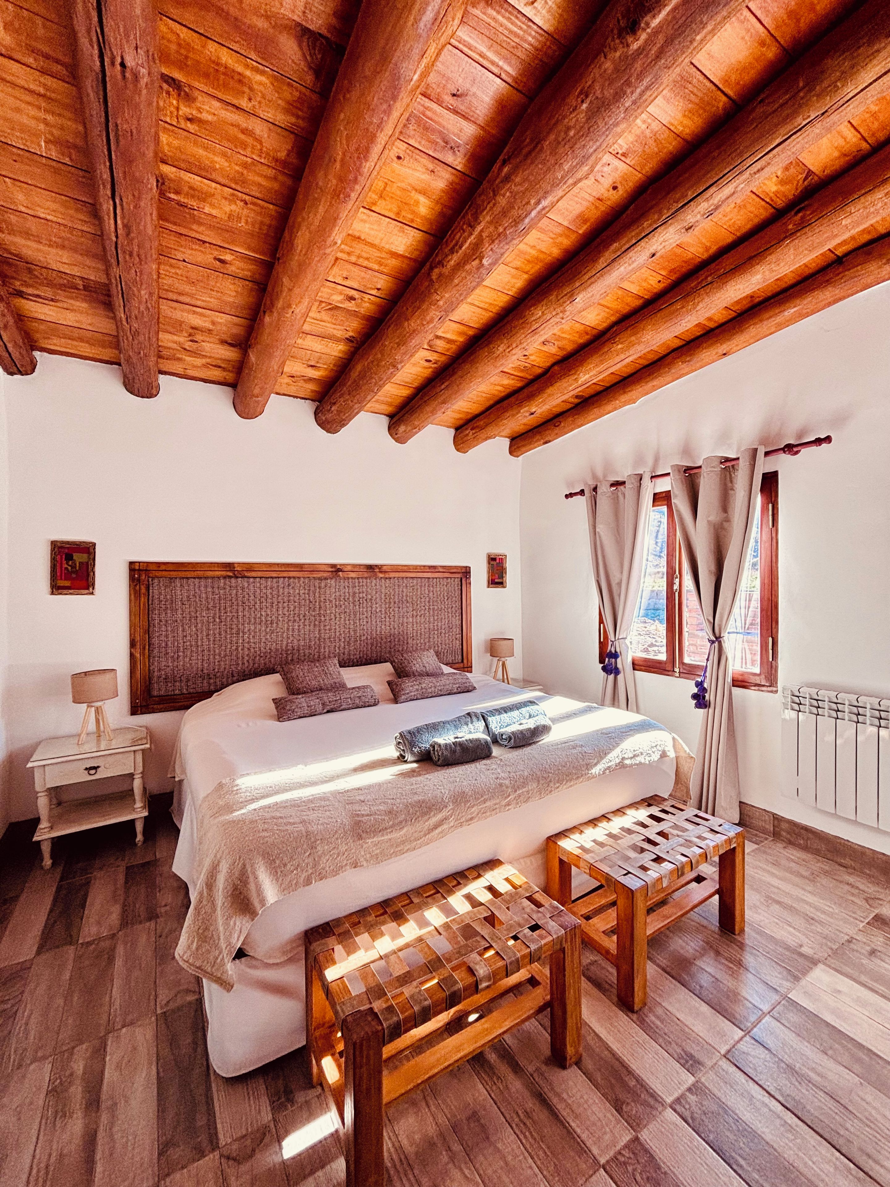 Un dormitorio con techo de madera, una cama grande, una mesita de noche, un banco y una ventana con cortinas.