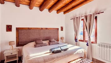 Un dormitorio con techo de madera, una cama grande, una mesita de noche, un banco y una ventana con cortinas.