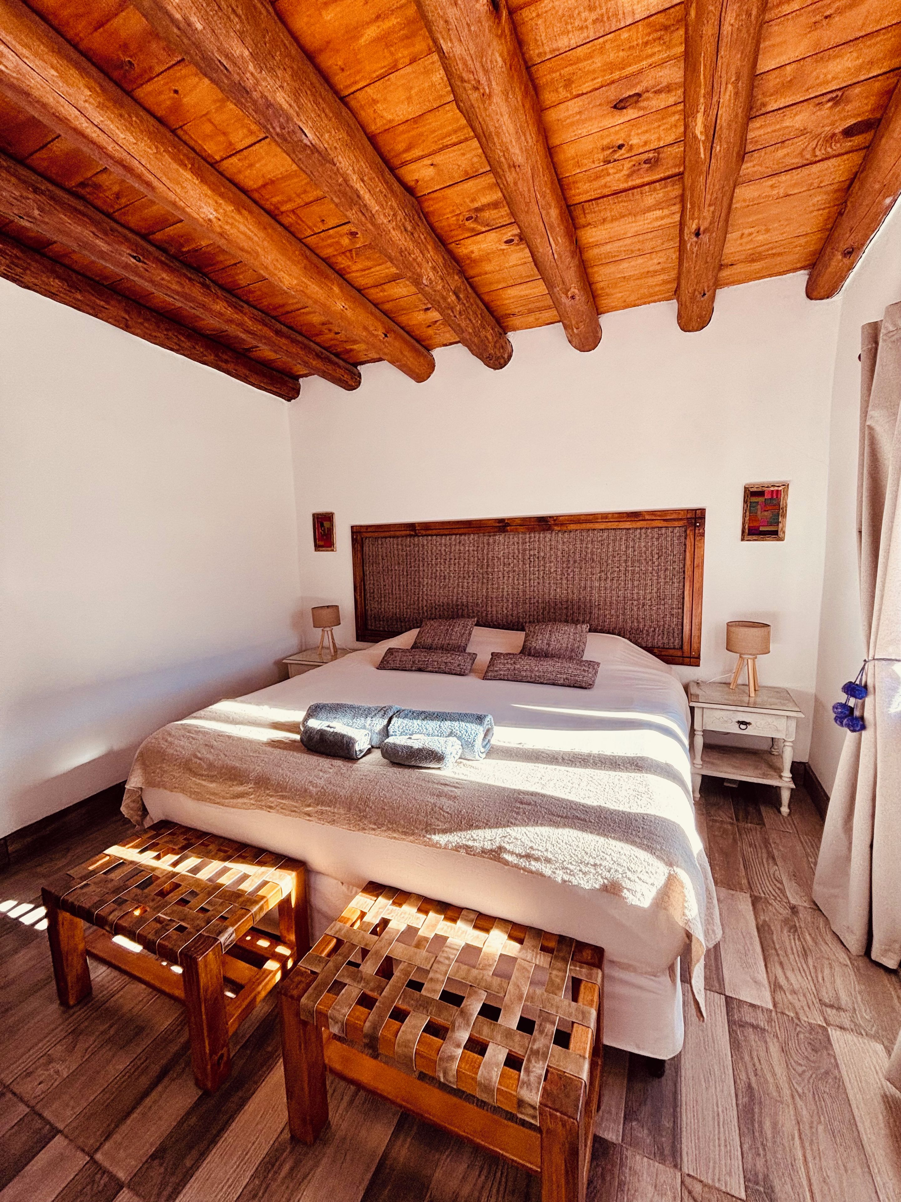 A bedroom with a wooden ceiling, a large bed with a headboard, two bedside tables, and a bench at the foot of the bed.