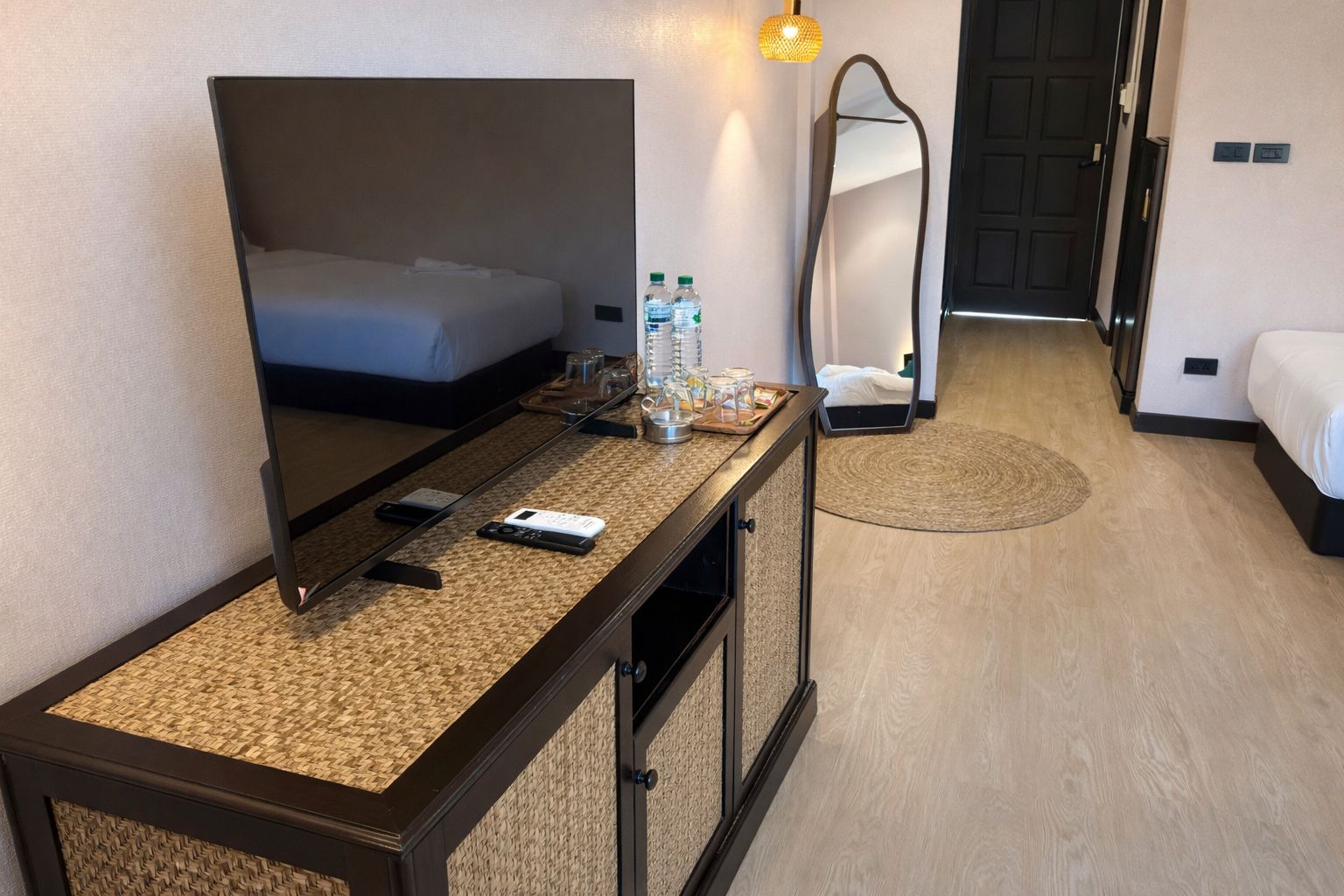 A hotel room with a bed, a bedside table with a mirror, a TV, and a water bottle.
