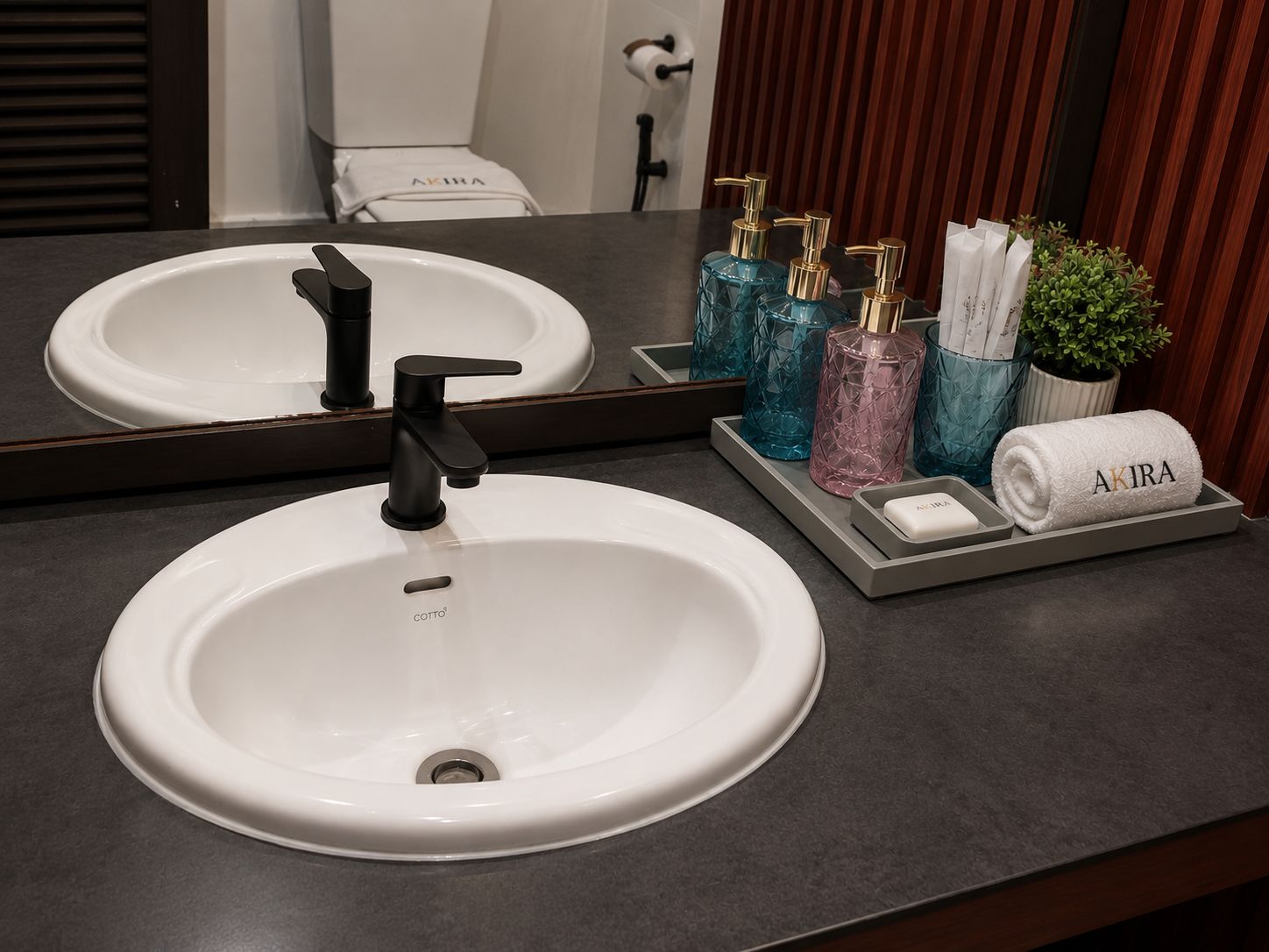 A bathroom with a white sink, black faucet, and a mirror reflecting the room. There are decorative bottles and a towel with 'AKIRA' on it.