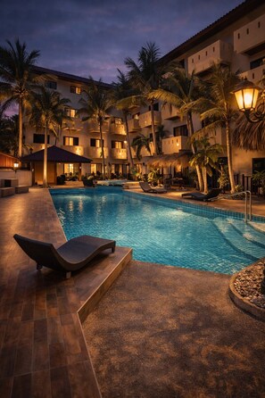 Una piscina con sillas de descanso, palmeras y un edificio con balcones al fondo.