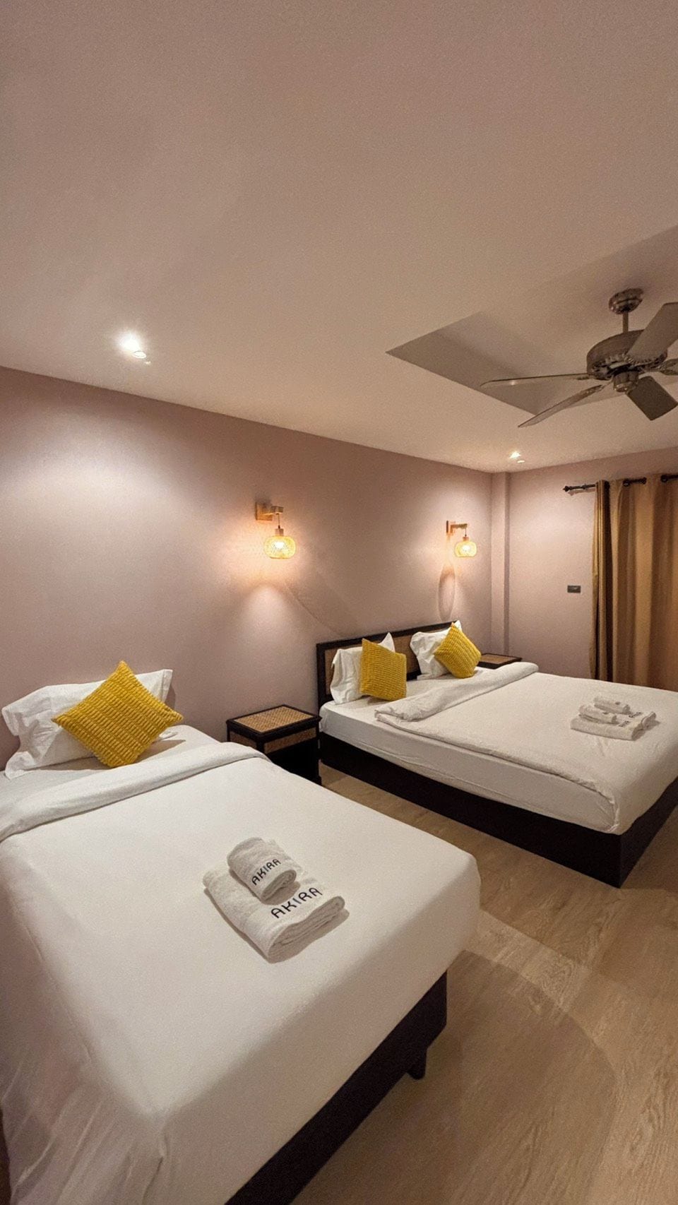 A hotel room with two beds, each with white linens and yellow pillows, a ceiling fan, and wall-mounted lights.