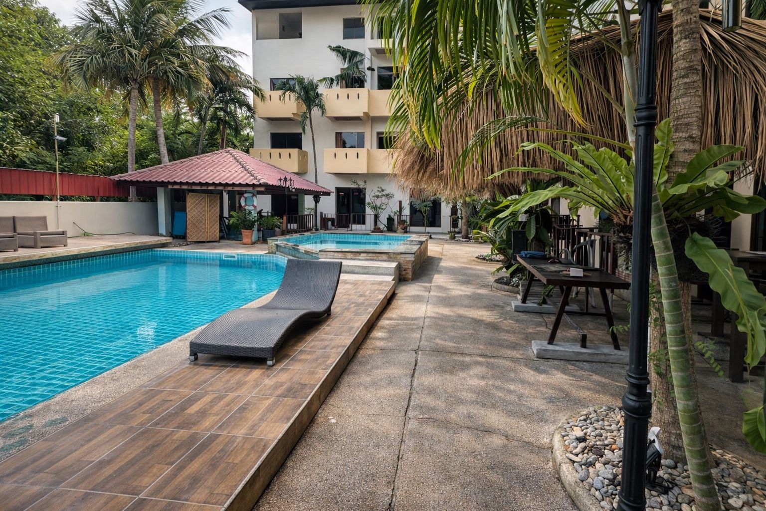 A swimming pool with lounge chairs, a thatched-roof structure, and a multi-story building with balconies.