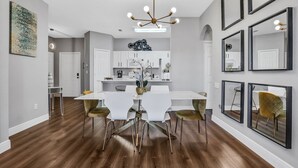 Ein moderner Essbereich mit einem weißen Tisch und Stühlen, einer Chandelierleuchtre und einer Küche im Hintergrund.