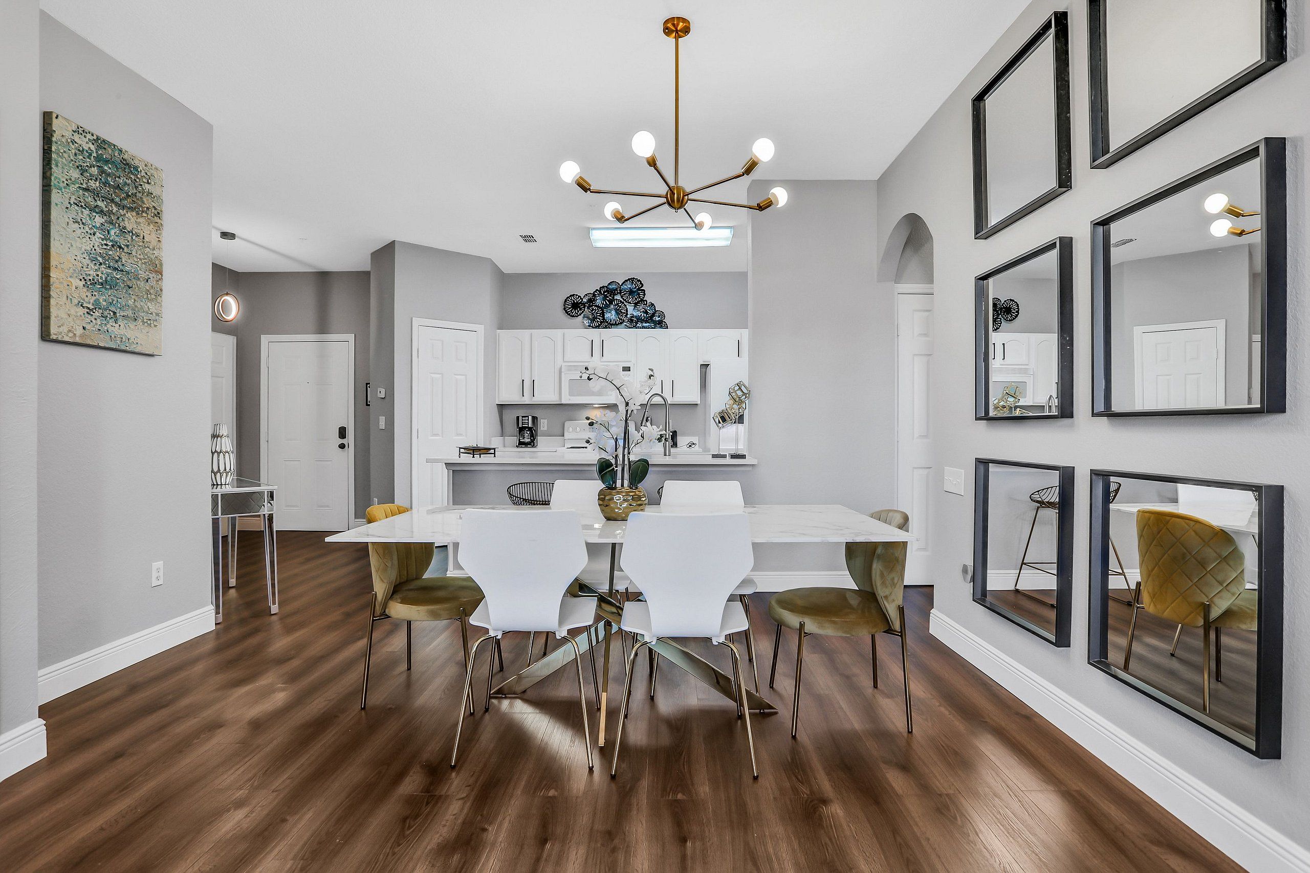 A modern dining area with a white table and chairs, a chandelier, and a kitchen in the background.