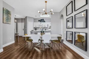 Un'area pranzo moderna con tavolo e sedie bianche, un lampadario e una cucina sullo sfondo.