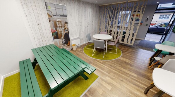 Un espace intérieur moderne doté d’un plancher en bois, d’une table de pique-nique verte, d’une table ronde avec des chaises et d’une cloison blanche ornée d’un motif d’arbre.