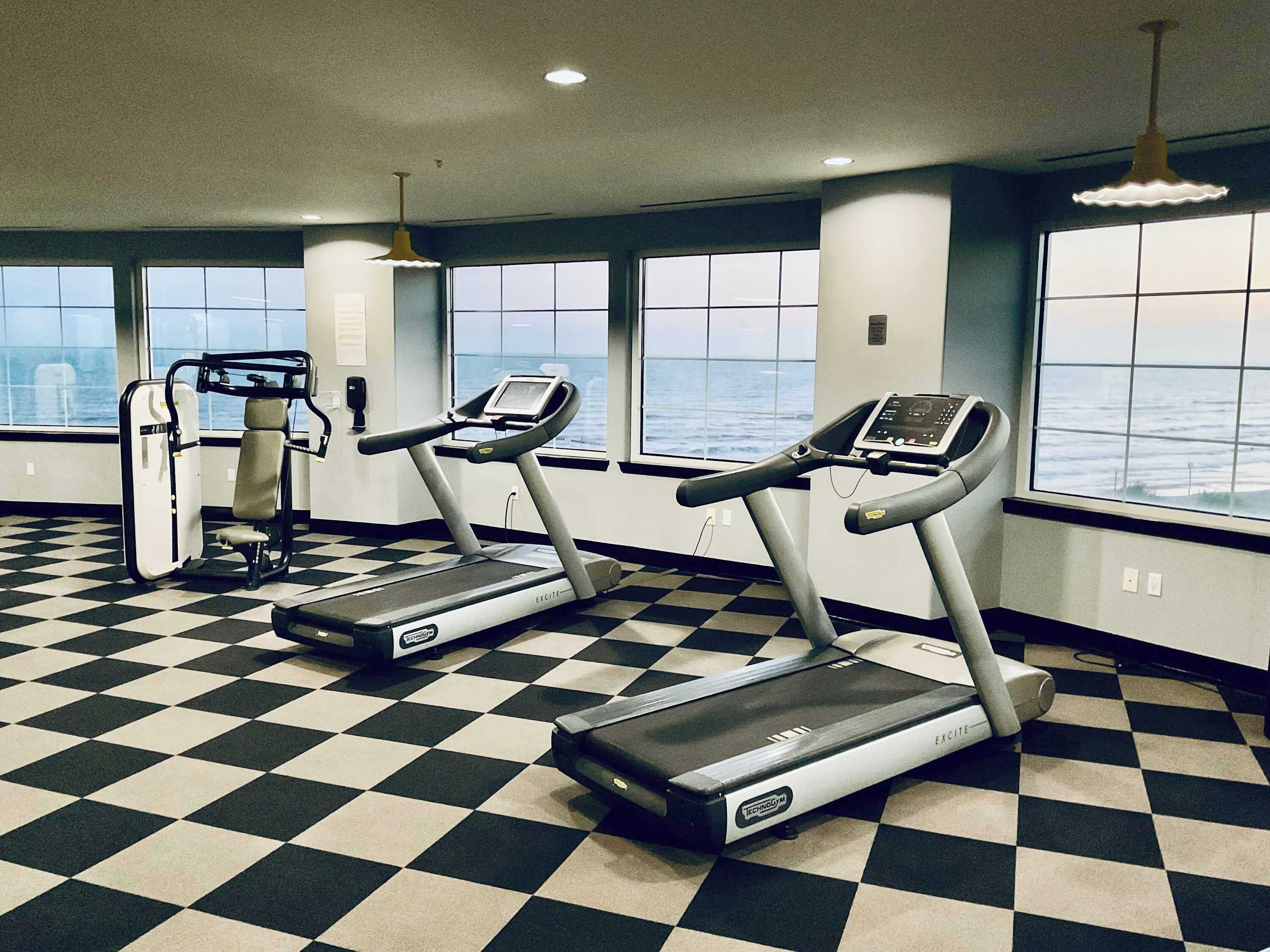 Une salle de sport équipée de deux tapis de course, d’une machine de musculation et de grandes fenêtres donnant sur un plan d’eau.
