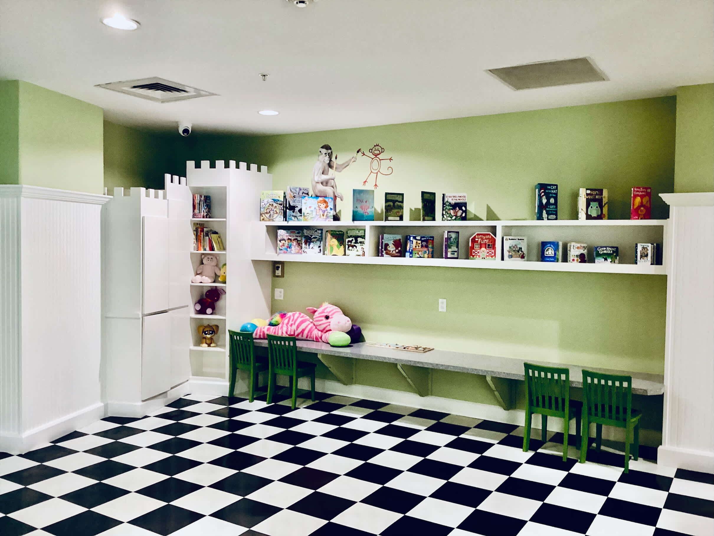 Una sala de juegos infantil con piso a cuadros, paredes verdes y estanterías blancas llenas de libros y juguetes.