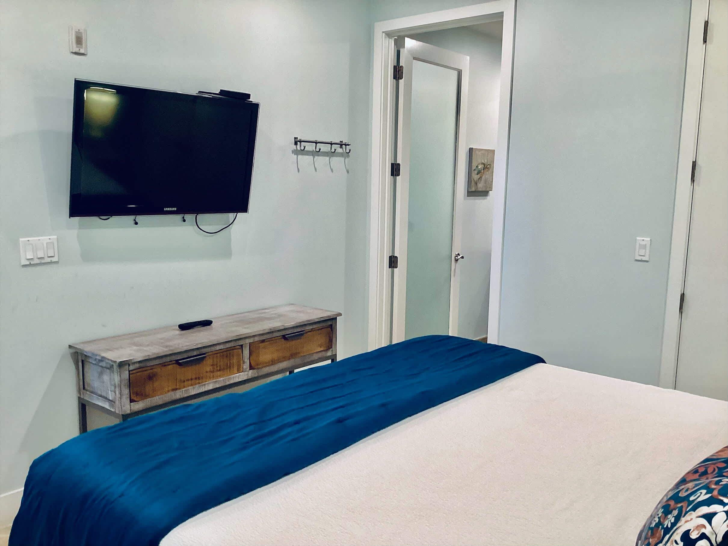 A bedroom with a bed, a television mounted on the wall, a wooden dresser, and a door leading to another room.