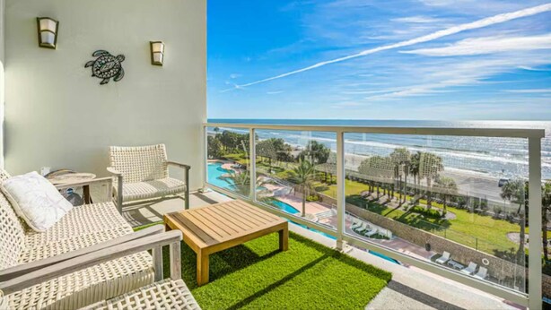A balcony with wicker furniture, a wooden table, and a view of a pool and ocean. - Diamond Beach Resort-escape #516 (Galveston)
