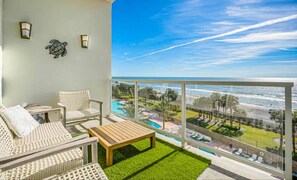 A balcony with wicker furniture, a wooden table, and a view of a pool and ocean. - Diamond Beach Resort-escape #516 (Galveston)