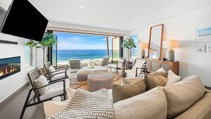 A modern living room with a large window overlooking a beach, a flat-screen TV mounted on the wall, and a fireplace. - Featured on HGTV "Christina On The Coast" - Stunning Ocean Views, Spa! (San Diego)