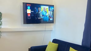 Un soggiorno moderno con un divano blu, due cuscini verdi e un tavolino da caffè bianco con gambe dorate. Una TV a schermo piatto montata a parete mostra l'interfaccia di Netflix.