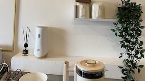 Una cucina con mobili bianchi, un piano di lavoro bianco, una macchina per il caffè, un bollitore, una pianta sospesa e una sveglia appesa al muro.