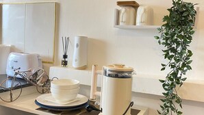 Un piano di lavoro in cucina con una macchina da caffè, un tostapane e un frigorifero. C'è una sveglia appesa al muro e uno scaffale con oggetti decorativi.