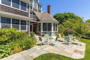 A house with a covered patio, a brick chimney, and a garden with green chairs. - Town & Tide Home for 8 (Kennebunkport)
