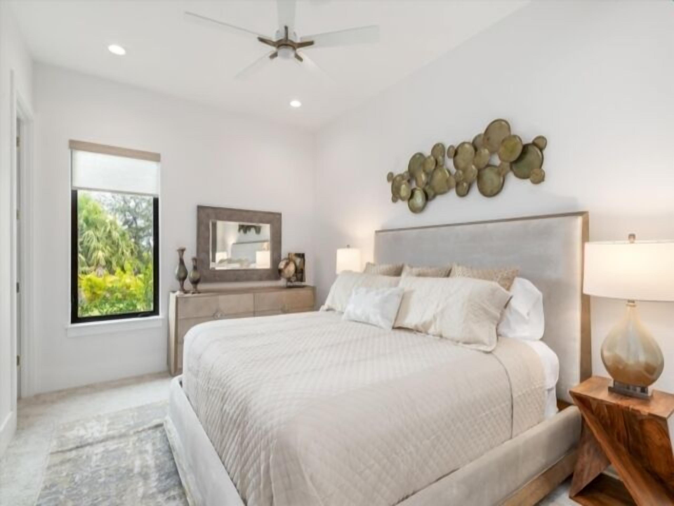 A bedroom with a large bed, a ceiling fan, a nightstand, a lamp, and a window with a view of greenery.