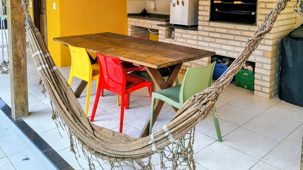 Un hamac, une table en bois et des chaises colorées dans une pièce lumineuse, avec un four en briques et un sol carrelé.