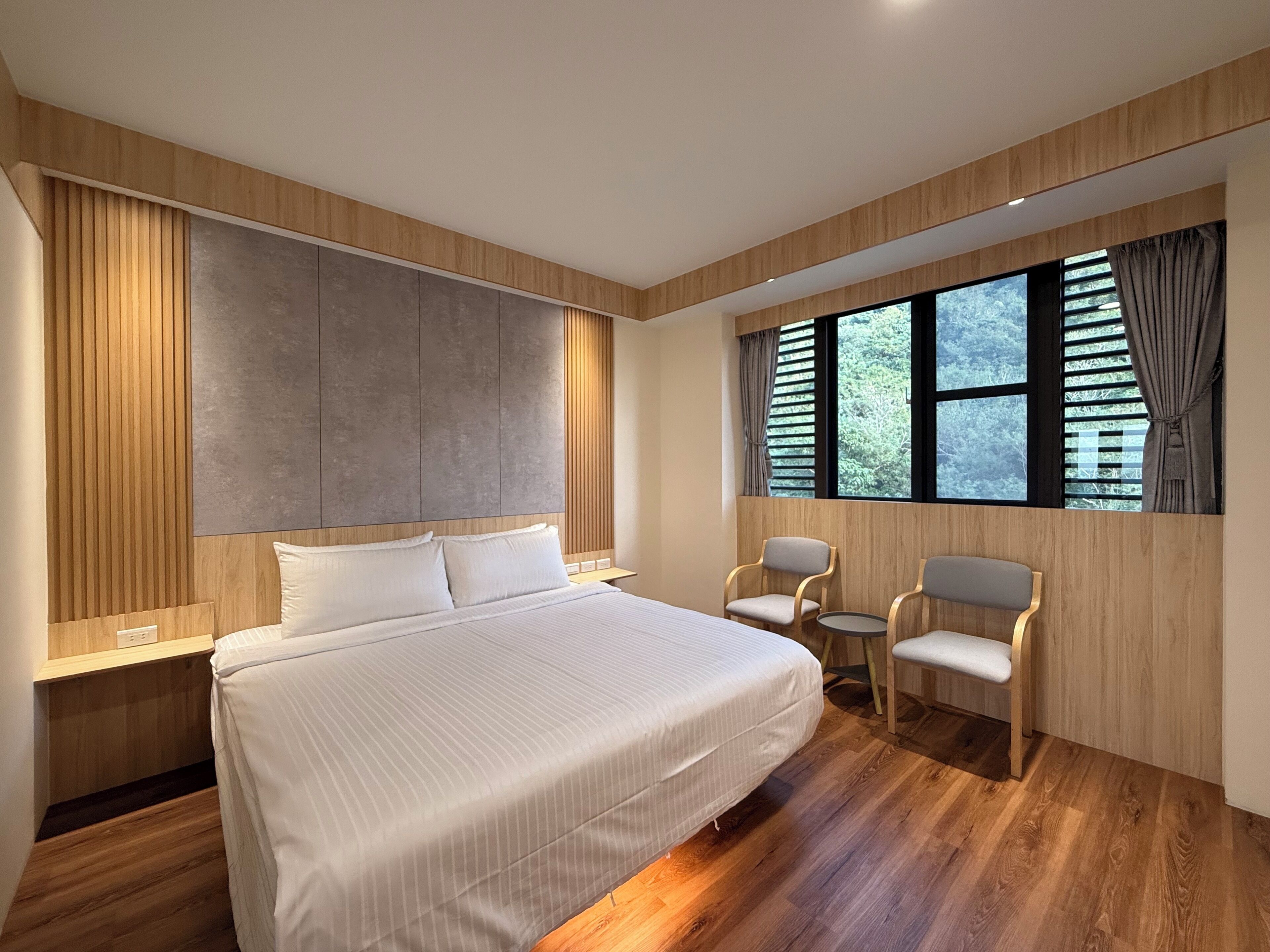 A modern hotel room with a large bed, two chairs, a window with blinds, and a wooden panel wall.