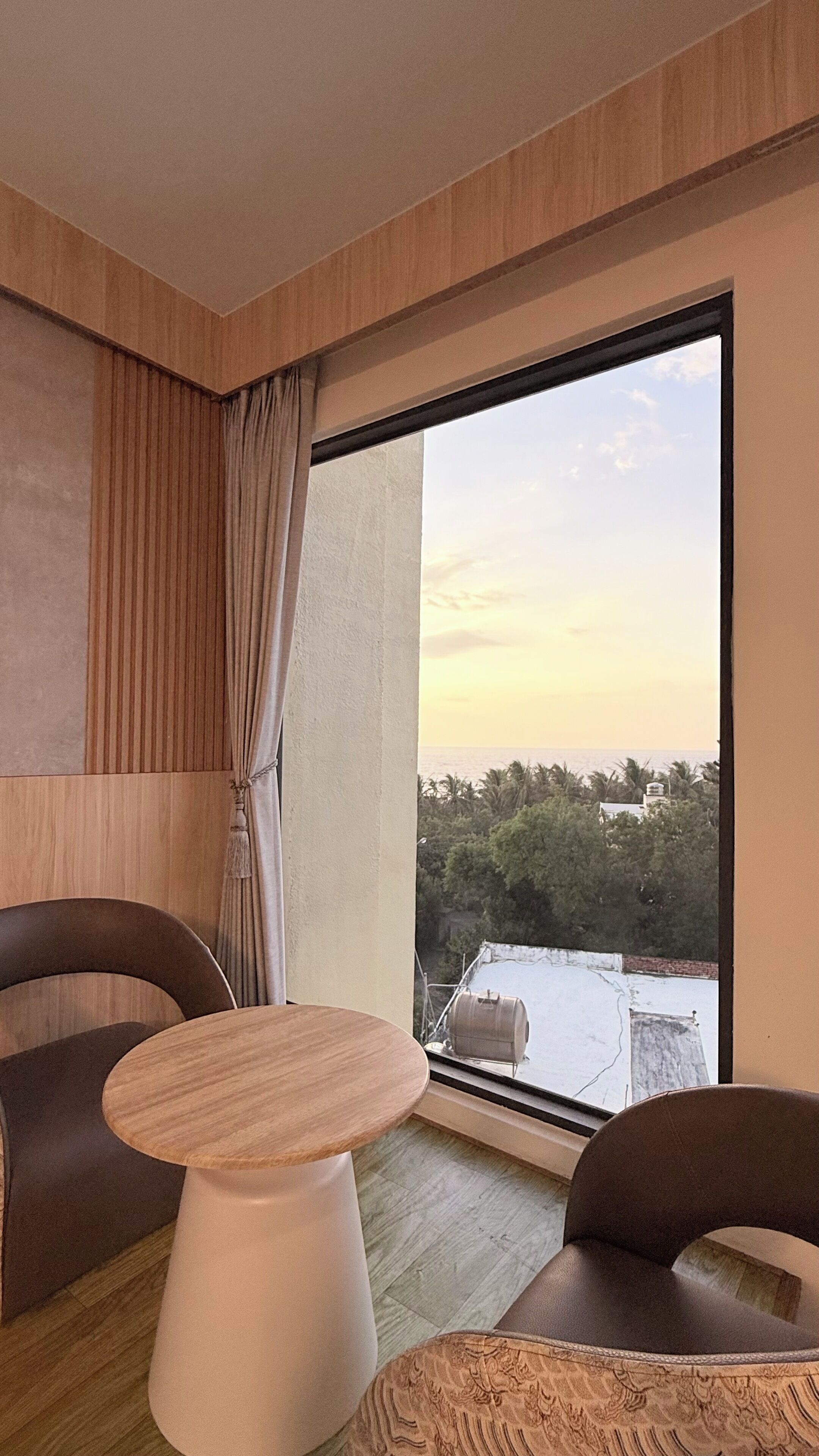 A modern room with a large window, a round table, two chairs, and a view of the sea and buildings.