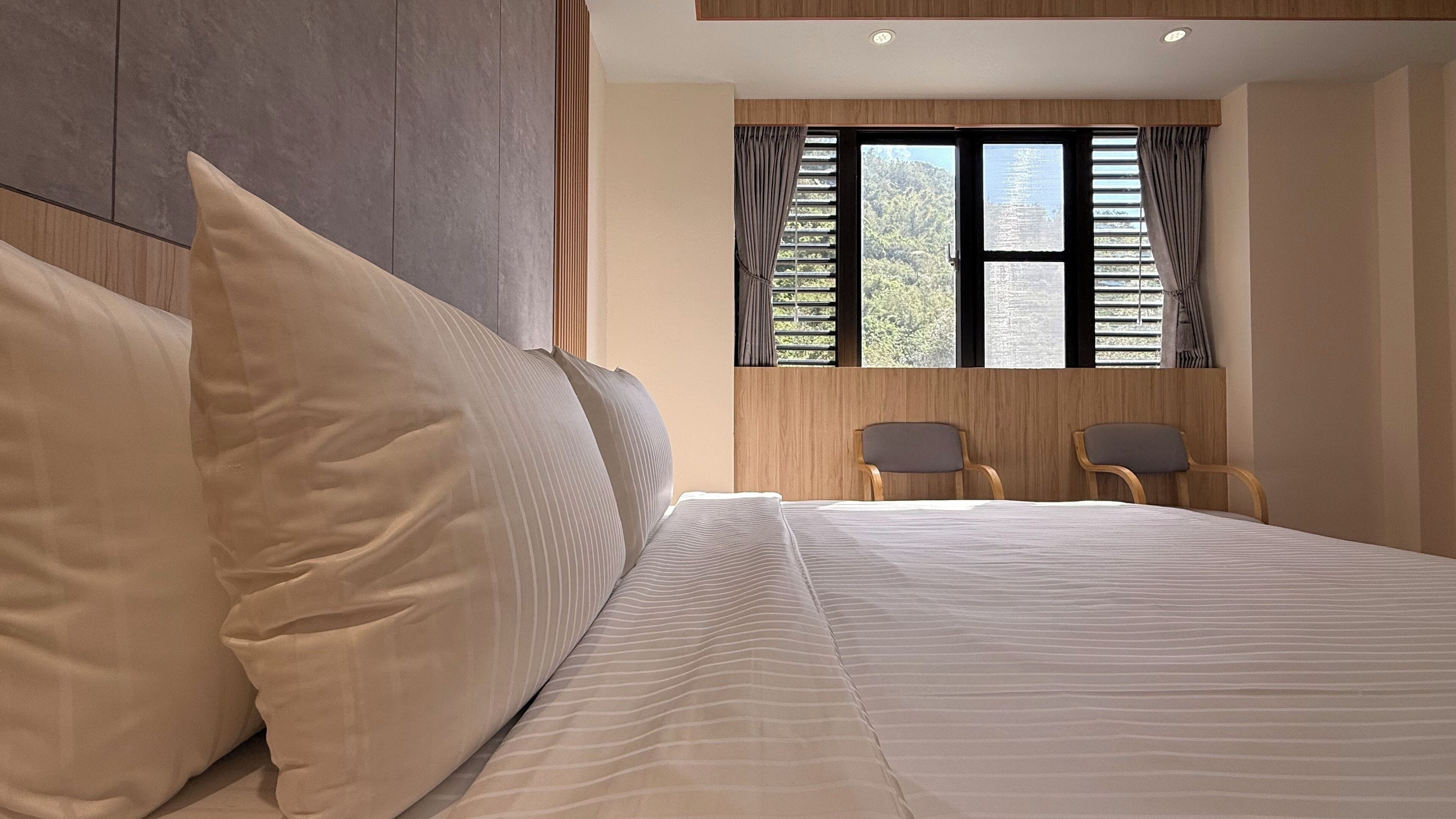 Ein Schlafzimmer mit einem großen Bett, zwei Stühlen, einem Fenster mit Vorhängen und Blick auf einen Wald.
