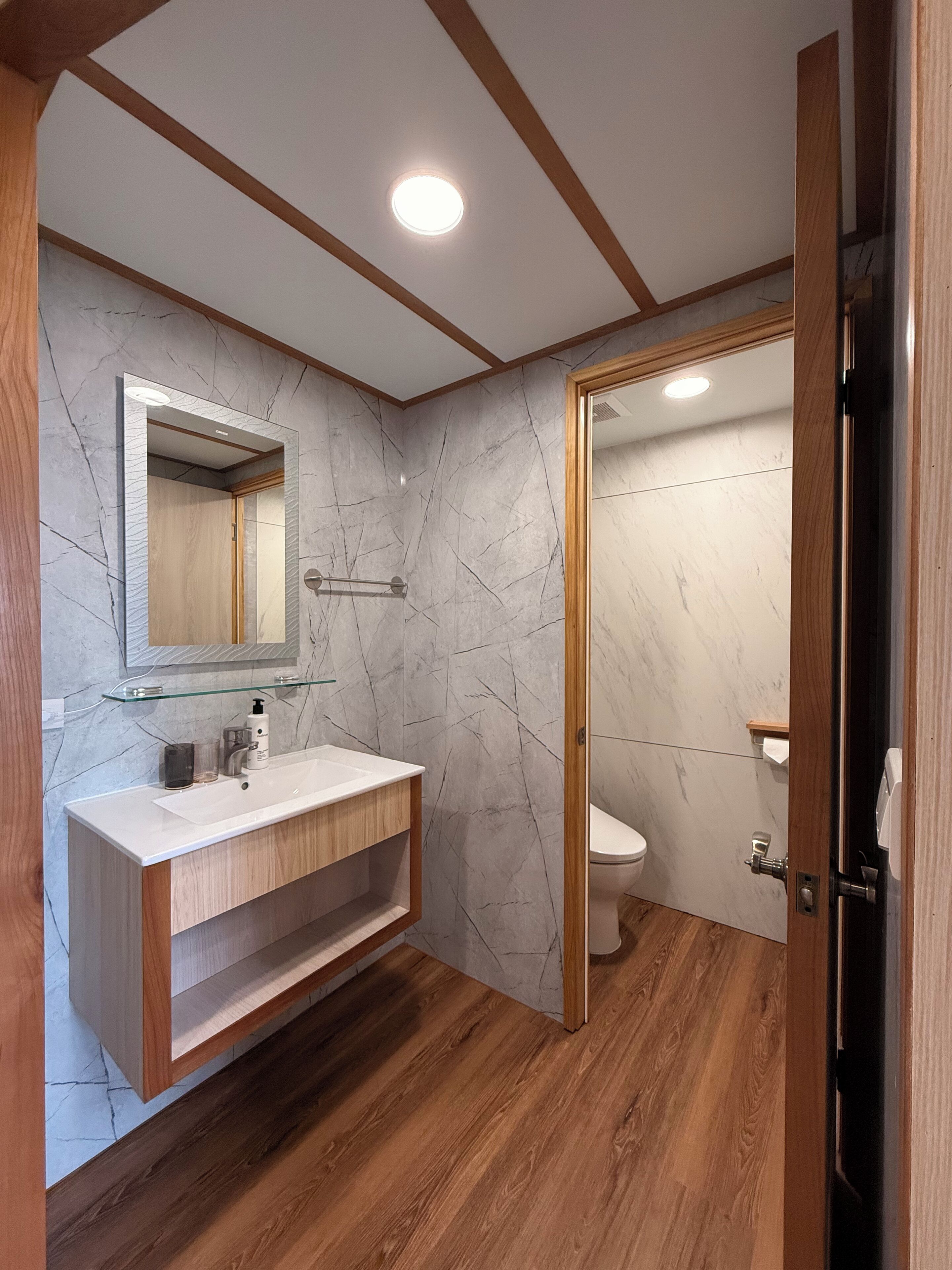 A modern bathroom with a marble wall, wooden vanity, and a toilet visible through an open door.