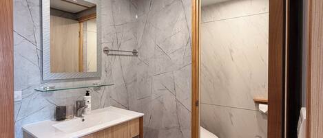 A modern bathroom with a marble wall, wooden vanity, and a toilet visible through an open door.