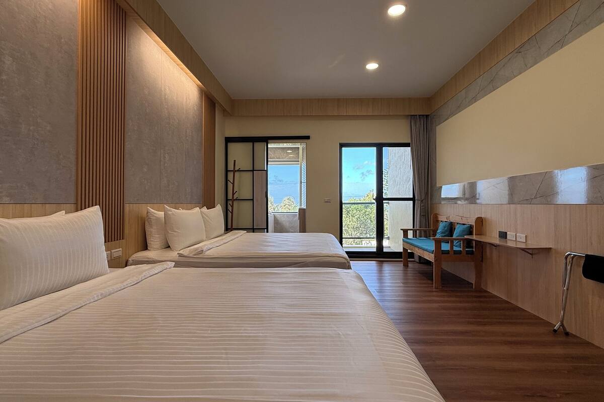 A hotel room with two beds, a wooden bench, a view of the outdoors, and a large window.