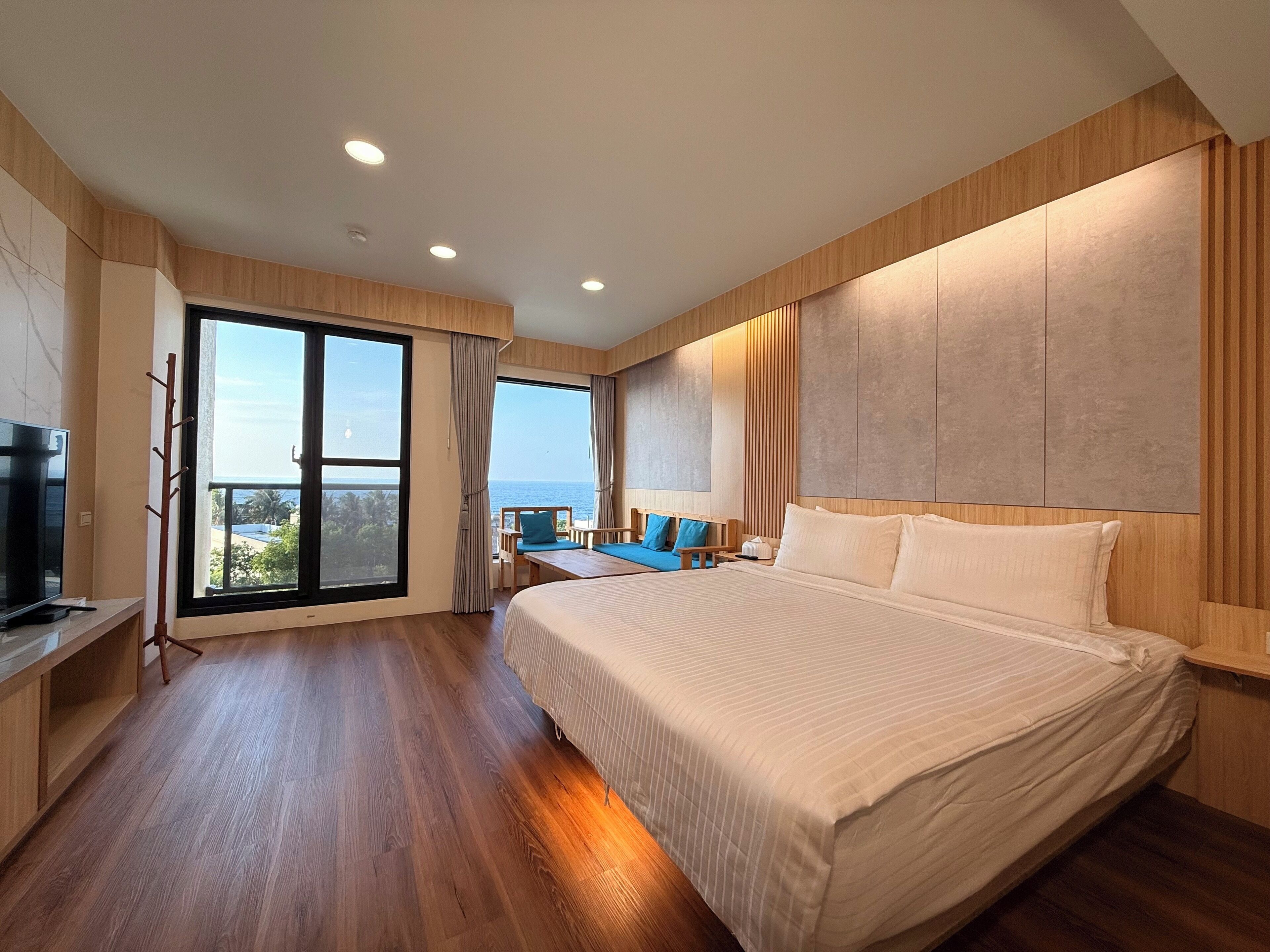 A modern hotel room with a large bed, a TV, a balcony with a view of the sea, and wooden flooring.