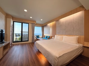 A modern hotel room with a large bed, a TV, a balcony with a view of the sea, and wooden flooring.