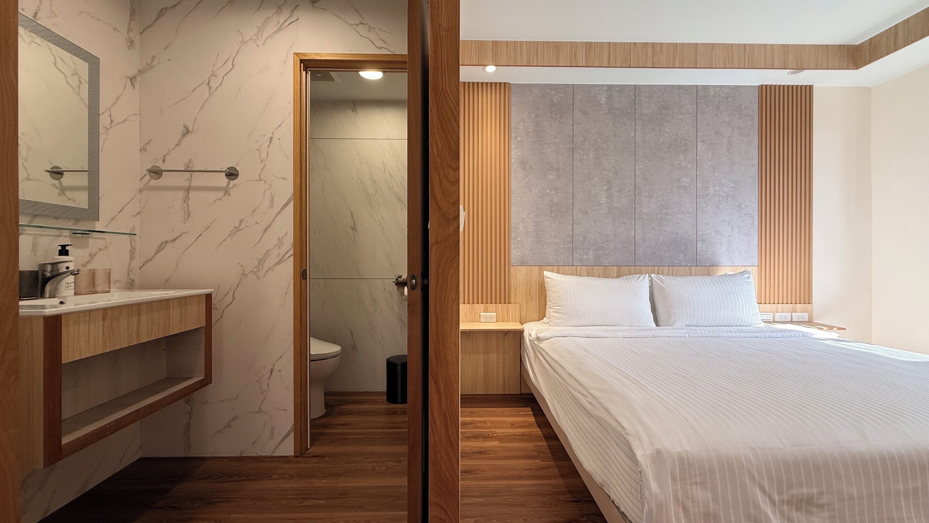 A modern hotel room with a wooden floor, a marble bathroom wall, a wooden headboard, and a large bed.