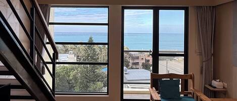 Una habitación amplia con piso de madera, una escalera, un ventanal con vista al mar y una cama con un almohadón azul.