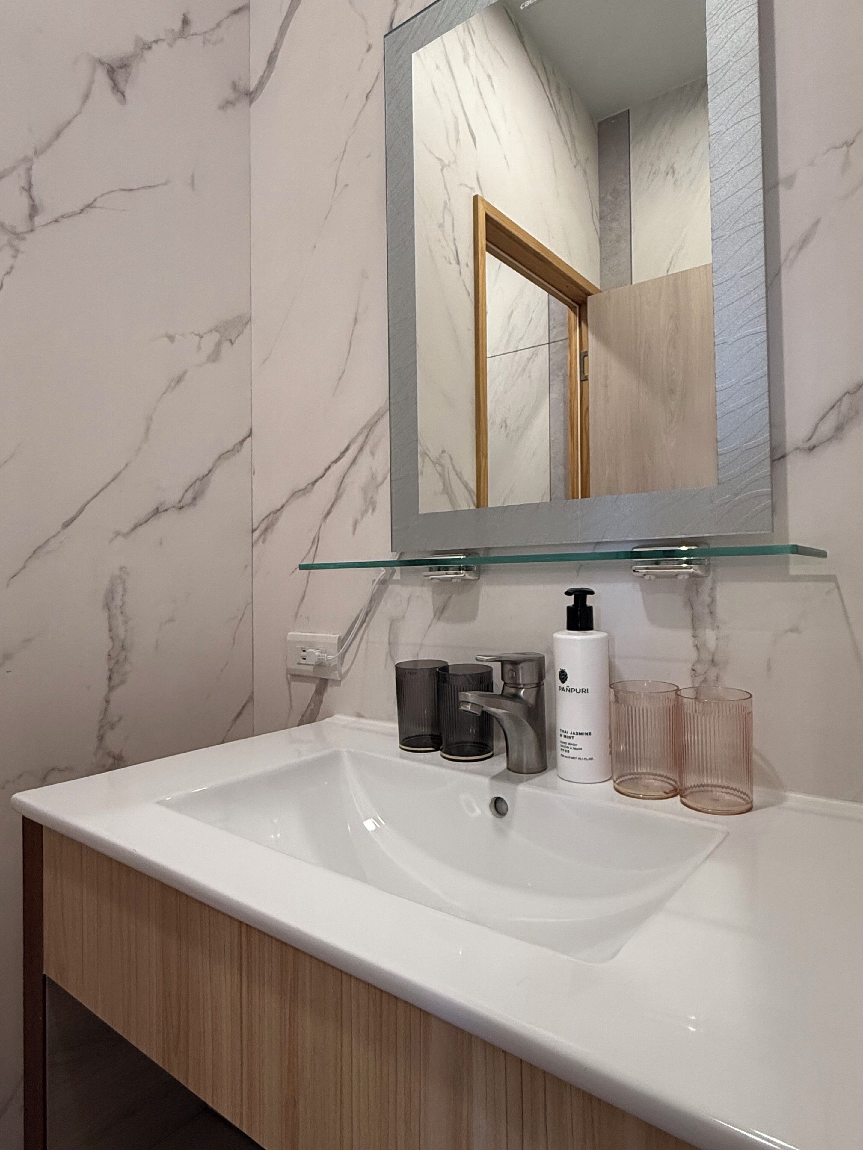 Un baño con lavamanos de mármol blanco, un espejo con marco de vidrio y un mueble de tocador de madera.