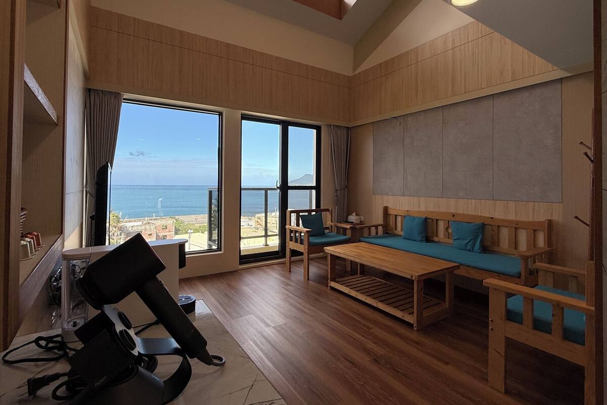A spacious room with a large window offering a view of the sea, a wooden sofa with teal cushions, a wooden coffee table, and a desk with a computer and headphones.